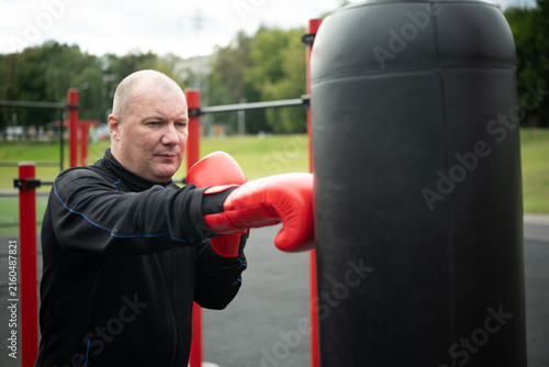 Fotografía Mature caucasian man in red boxing gloves punching heavy bag at outdoor gym, strength training, cardio workout and active aging concept
