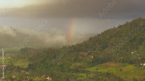 Aerial view of Sidemen Rice Terrace with a rainbow over lush green hills and tropical vegetation in Kecamatan Sidemen, Bali, Indonesia.