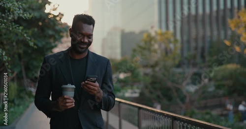 African American businessman checks his smartphone while holding takeaway coffee and walking outside the office, blending a city work break with mobile business.