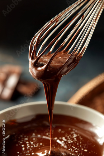 Rich melted dark chocolate dripping from a metal wire whisk into a glossy bowl, creating a perfect smooth stream, ideal for baking and luxury desserts
