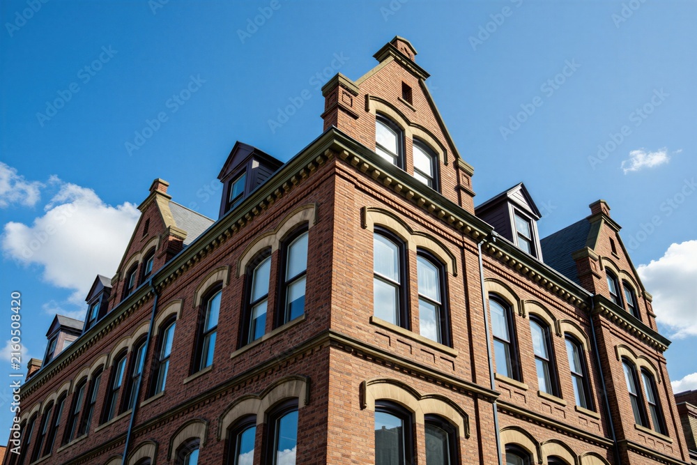 Fototapeta premium Low angle view of classic red brick building with architectural details against blue sky. Multi story construction features historical masonry facades, large arched windows with attic dormers.