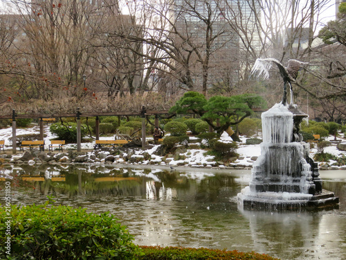 View of a frozen fountain with a crane statue surrounded by snow-covered grounds, bare trees, and park benches in Hibiya Park, Tokyo, Tokyo, Japan.