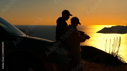Couple drinking coffee during road trip at sunset. Silhouettes of a man and a woman drinking hot beverages from mugs while leaning against a car, enjoying the stunning ocean sunset view