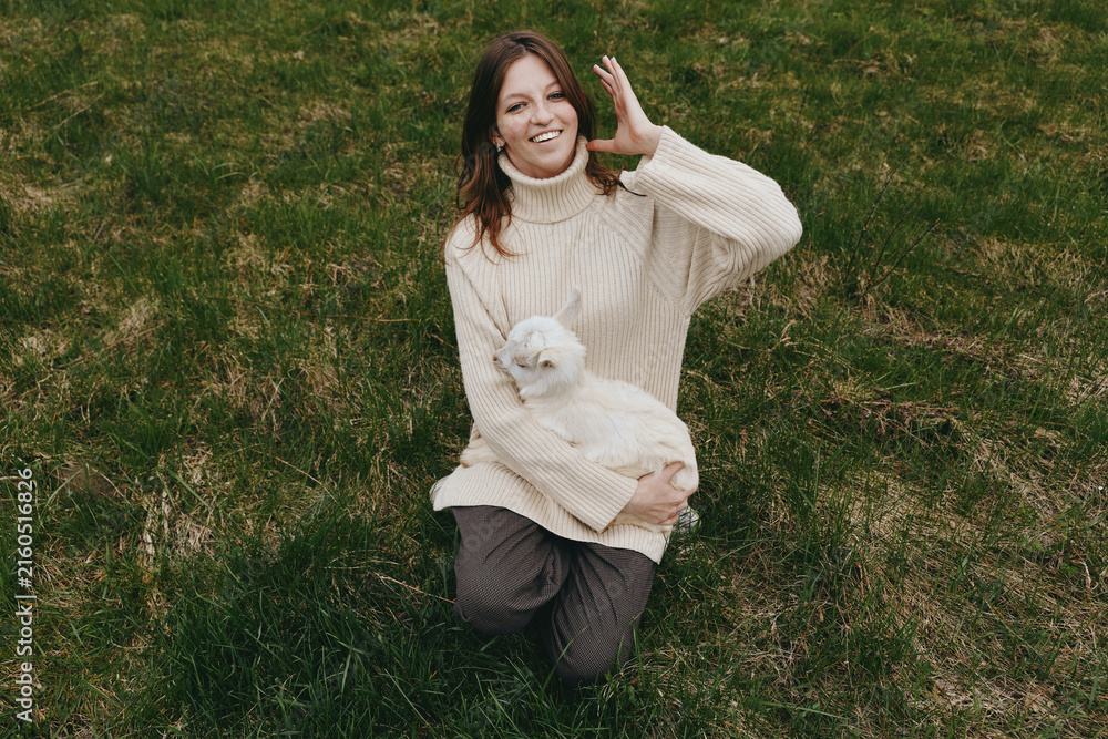 Fototapeta premium Cheerful woman outdoors holding a goat kid in a sunlit field, cozy sweater, relaxed smile, gentle breeze, natural colors, lush grass, rural lifestyle moment