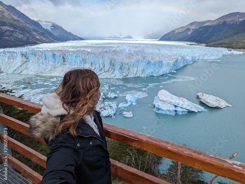 Obraz na plátne Dramatic high-angle view of a woman at a railing, back to camera, observing Perito Moreno Glacier with floating icebergs and a double rainbow above, Patagonia, Argentina