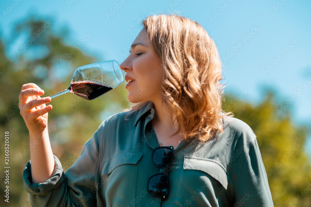 Naklejka premium Woman smelling red wine in glass outdoors on sunny day, in green shirt and sunglasses. Wine tasting experience and relaxing weekend leisure.