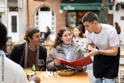 Obraz na płótnie On open area cafe terrace, guy waiter communicates with girl and her boyfriend customers, answer questions about composition of dish of day