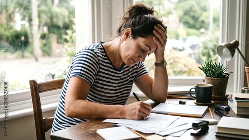 Tired young woman sitting at a table with a notebook, feeling stressed and overwhelmed while studying or working at home.