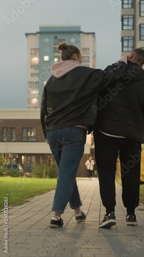 Woman and man stroll side by side along pathway. Footwear consists of shoes and sneaker on both subjects. Environment shows paved path lit windows on one building to left