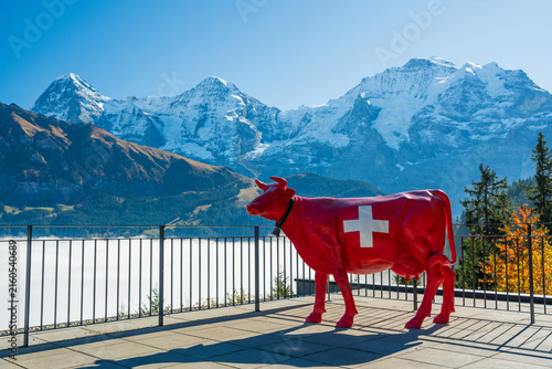Red cow statue with Swiss cross overlooking sea of clouds and snow-capped Eiger, Monch and Jungfrau mountains of the Alps. Murren, Canton of Bern, Switzerland.
