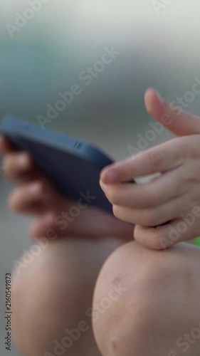 Close-up of a boy playing on a smartphone outdoors. Focus on hands holding the phone resting on bare knees against a blurred natural background.
