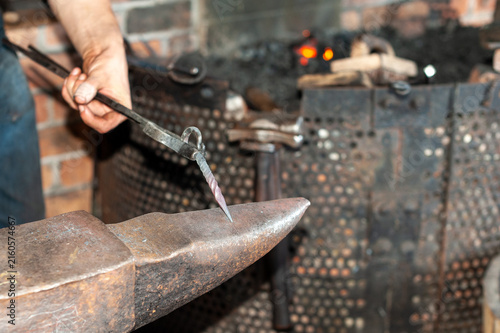 Принти на полотні A forger wearing blue jeans and a T-shirt holds fire tongs and a twisted metal bolt on a vintage anvil