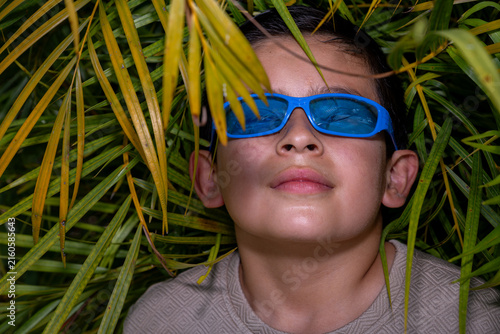 Young boy in blue sunglasses experiencing a sense of wonder and curiosity while peeking through lush green and yellow palm leaves, enjoying a playful outdoor adventure in nature