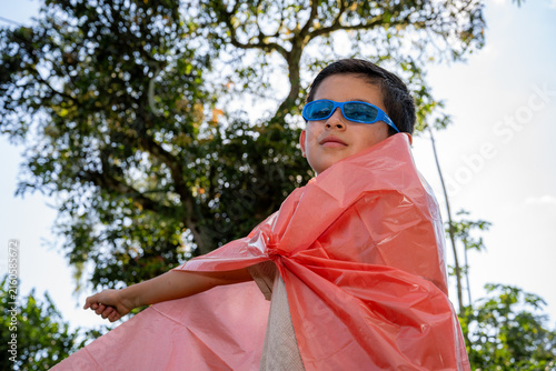 Young boy playing superhero outdoors, wearing blue sunglasses and a makeshift red cape made from plastic, imagining himself flying and saving the day