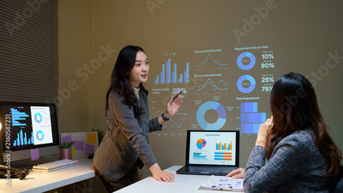 Confident Asian female professional presents marketing analytics charts on projected screen to colleague, ideal for business, teamwork, and corporate training concepts.