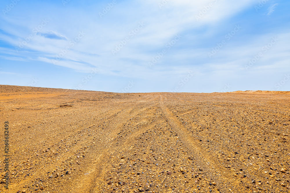 Obraz premium Barren gravel desert landscape in Dahaidao, Xinjiang, China, with tire track leading toward a low horizon beneath a blue sky, conveying remoteness and travel potential.