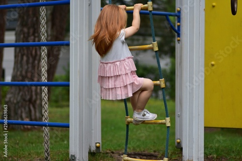 Fotografie Little red-haired girl climbing a rope ladder at an outdoor playground, reaching for the next rung while wearing a pink ruffled dress, enjoying active play in a green park setting