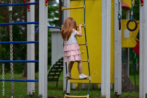 Fotografie Little red-haired girl climbing a rope ladder at an outdoor playground, reaching for the next rung while wearing a pink ruffled dress, enjoying active play in a green park setting