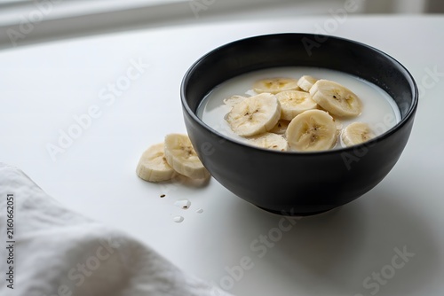 Minimalist breakfast featuring a black bowl of fresh sliced bananas and creamy milk on a bright white surface, capturing a clean, healthy, and modern morning mood.