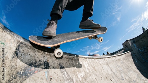 Low angle fisheye shot of a skateboarder performing an ollie jump over a concrete bowl ramp at a skatepark against a clear blue sky.