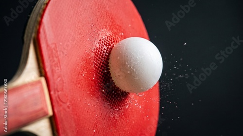 Macro extreme close-up of a white table tennis ball impacting a red ping pong paddle with spin dust against a dark background.