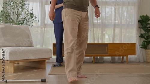 Older adult walking barefoot with caregiver bright living room, cautious movement and rehabilitation with walking cane, medical care, wooden furniture, plants, soft curtains, natural daylight