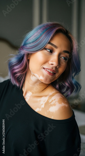 Smiling young asian woman with colorful hair and vitiligo looking over shoulder wearing black top indoors