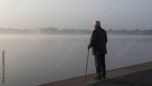 Senior Man with Cane Stands by Calm Misty Water at Dawn