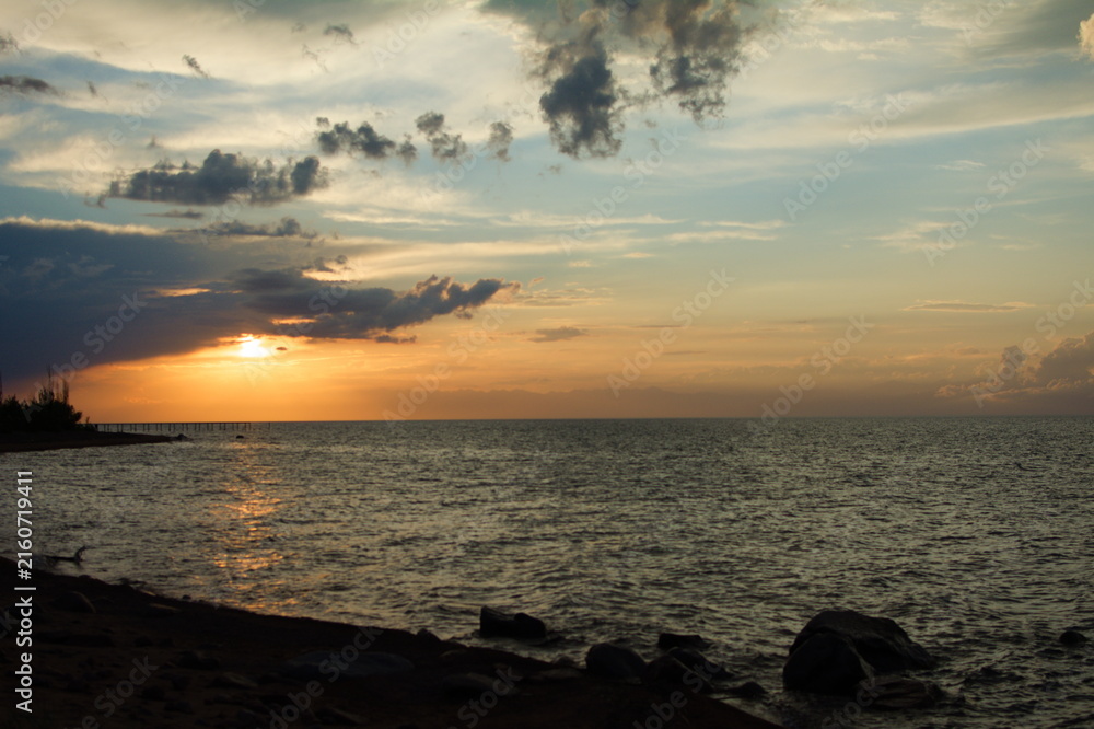 Obraz premium Tranquil seaside sunset with dark silhouettes of rocks and trees on the shore, vibrant orange and blue hues filling the sky beneath scattered clouds, reflecting on the calm ocean water.