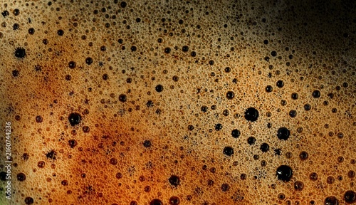 Rich amber beer foam bubbles gently against a dark backdrop. Each bubble holds light, catching warm tones of golden brown. The surface glistens with texture and depth, inviting close inspection