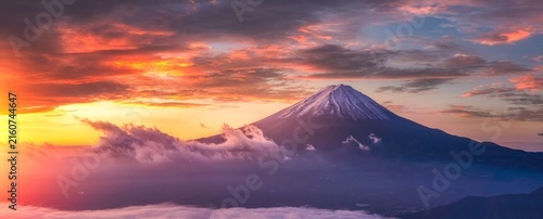 Golden sun kisses Mount Fuji's snowy peak. Clouds swirl below, glowing with sunset hues. Sky blurs orange, purple, and deep blue. Mount Fuji stands still, majestic and serene
