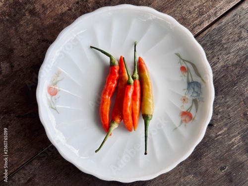Fotografie close-up, top-down photograph of five fresh, vibrant red and yellow-green bird's eye chilies with green stems, neatly arranged on a vintage white plate with a delicate floral pattern