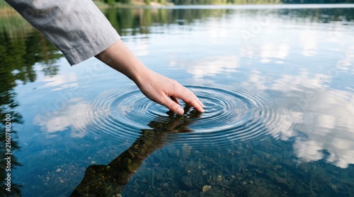 Hand touching calm lake water creating ripples, connection with nature, mindfulness and sensory experience, peaceful summer day, reflection of sky and clouds, therapeutic touch