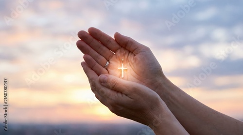 Hands holding glowing cross necklace in prayer position against soft sunset sky, symbolizing faith, religion, spirituality, and divine light or blessing.