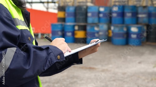 Checking on chemical hazardous safety checklist form, close-up at worker hand, with factory warehouse and metal barrel storage as background. Industrial safe work practice scene, selective focus.