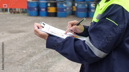 Checking on chemical hazardous safety checklist form, close-up at worker hand, with factory warehouse and metal barrel storage as background. Industrial safe work practice scene, selective focus.