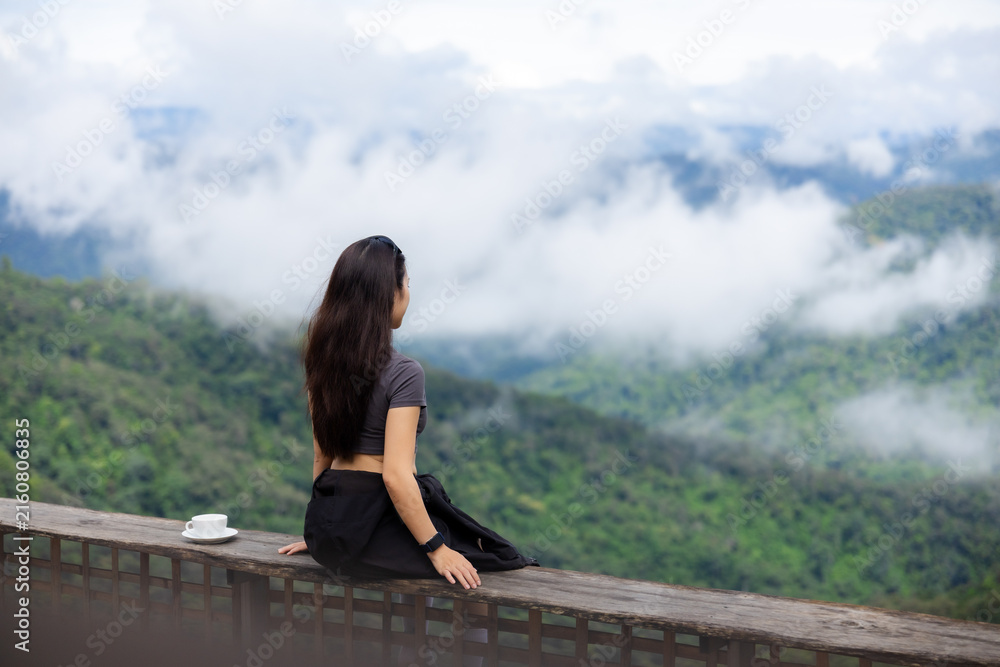Fototapeta premium Woman sitting on wooden balcony overlooking misty mountain valley, enjoying peaceful solitude among lush green hills and drifting cloud