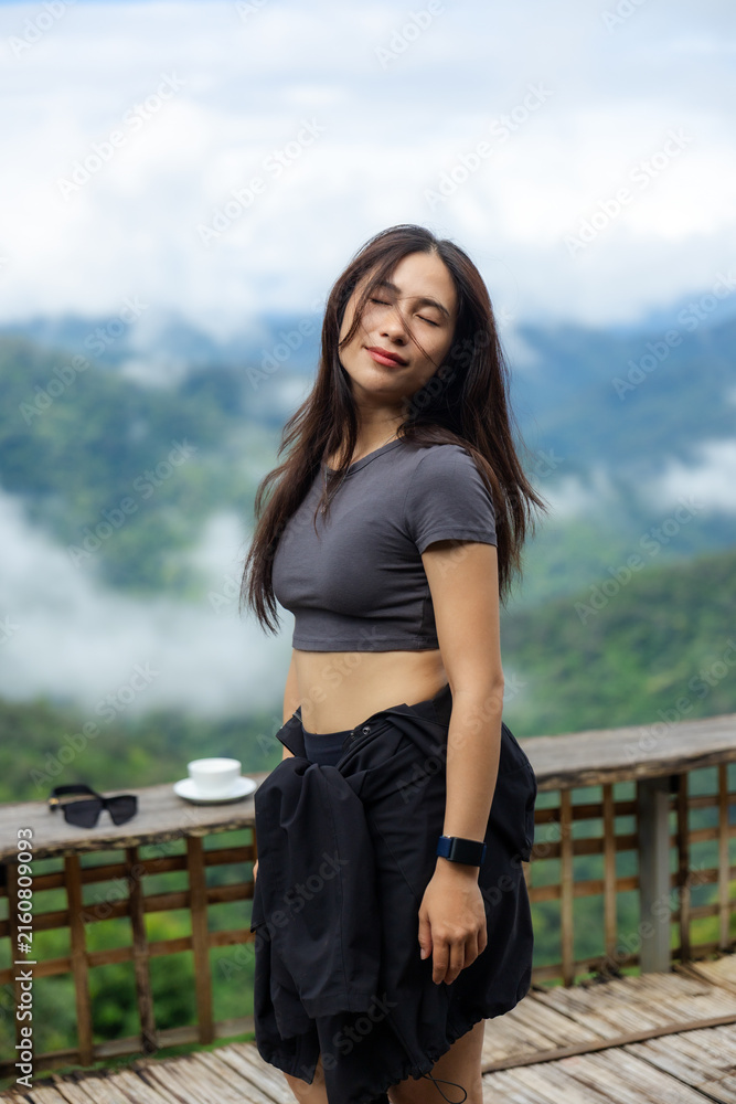 Fototapeta premium Young woman in dark shirt standing on wooden terrace above misty mountain valley, eyes closed with peaceful expression and wind moving her hair