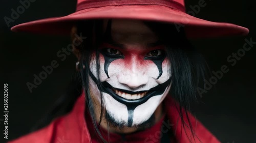 Close sinister smile clown under red wide brim hat with black eye paint for halloween poster background and horror banner design