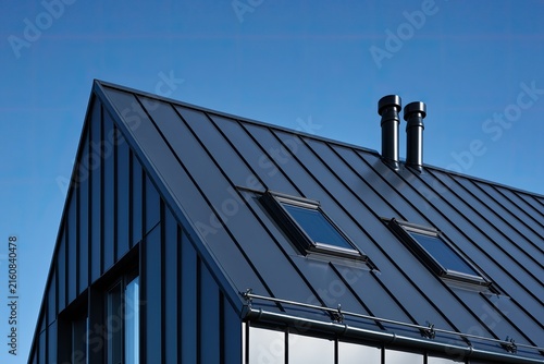Modern dark metal roof with gables, dormers, and chimneys under blue sky