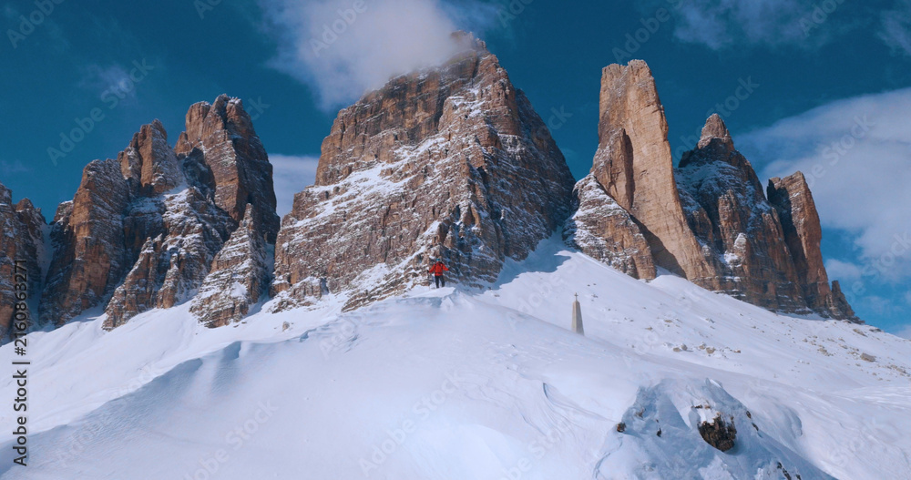 Obraz premium Ski mountaineer crossing a snowy ridge in the Dolomites with dramatic alpine peaks and blue sky in the background, capturing winter adventure and breathtaking mountain landscape.