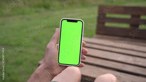 Hand holds phone with green screen outdoors on wooden bench. Person relaxed, seated, focused on device. Perfect for app demos or advertising mockups. Natural daylight, grassy background