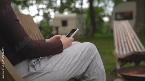 Person sits on wooden chair, focused on phone. Relaxing outdoors near campfire pit. Calm, peaceful atmosphere with green grass and trees. Ideal for travel, camping, or digital detox themes
