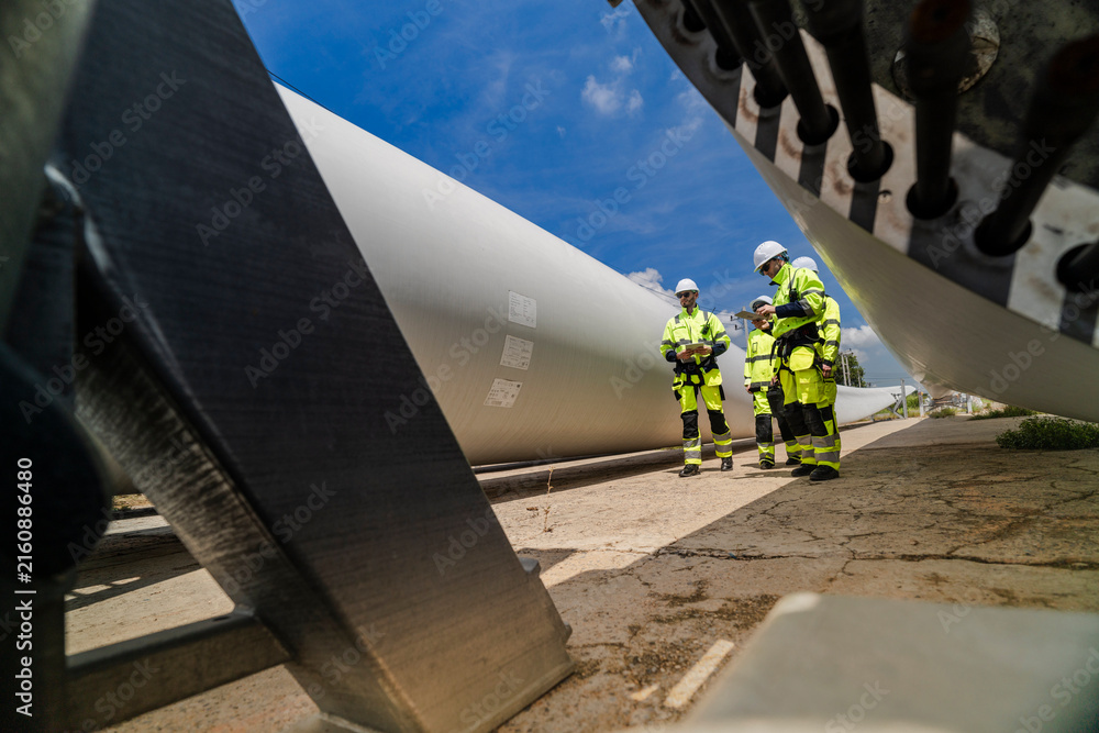Fototapeta premium Four technicians in yellow and navy coveralls stand in a loose group under the bolted root end of a turbine blade that is parked on its cradle in a component laydown yard