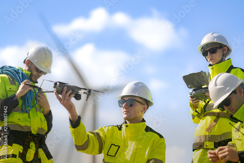 A clean-shaven technician holds a folded inspection drone up on his fingertips while a bearded colleague with a blue rope coil and two teammates with a controller look on