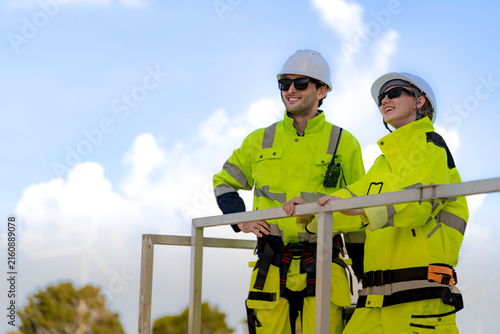 Two wind farm workers stand talking beside a galvanised steel access frame, one turned toward the other with the tubular rails running out of the right side of the frame