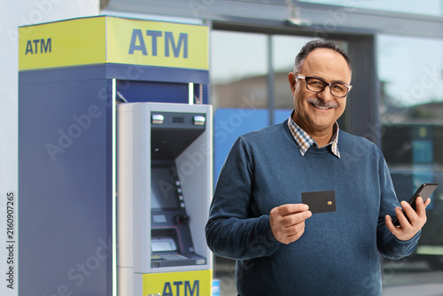 Fotografía Mature man holding a credit card and a mobile phone next to an ATM