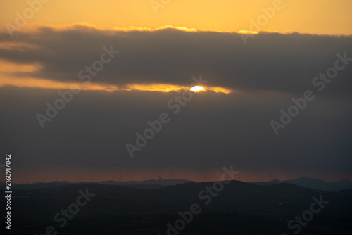 Foto Sun peeking through dark clouds during a serene sunset over distant hills