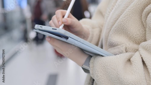 Close-up of hand holding tablet and stylus. Person wears fluffy beige coat. Background shows blurred people walking. Focus remains on digital writing action. Urban environment with soft lighting