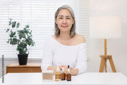 Photography An Asian mature woman with natural grey hair sits confidently at a table with skincare products